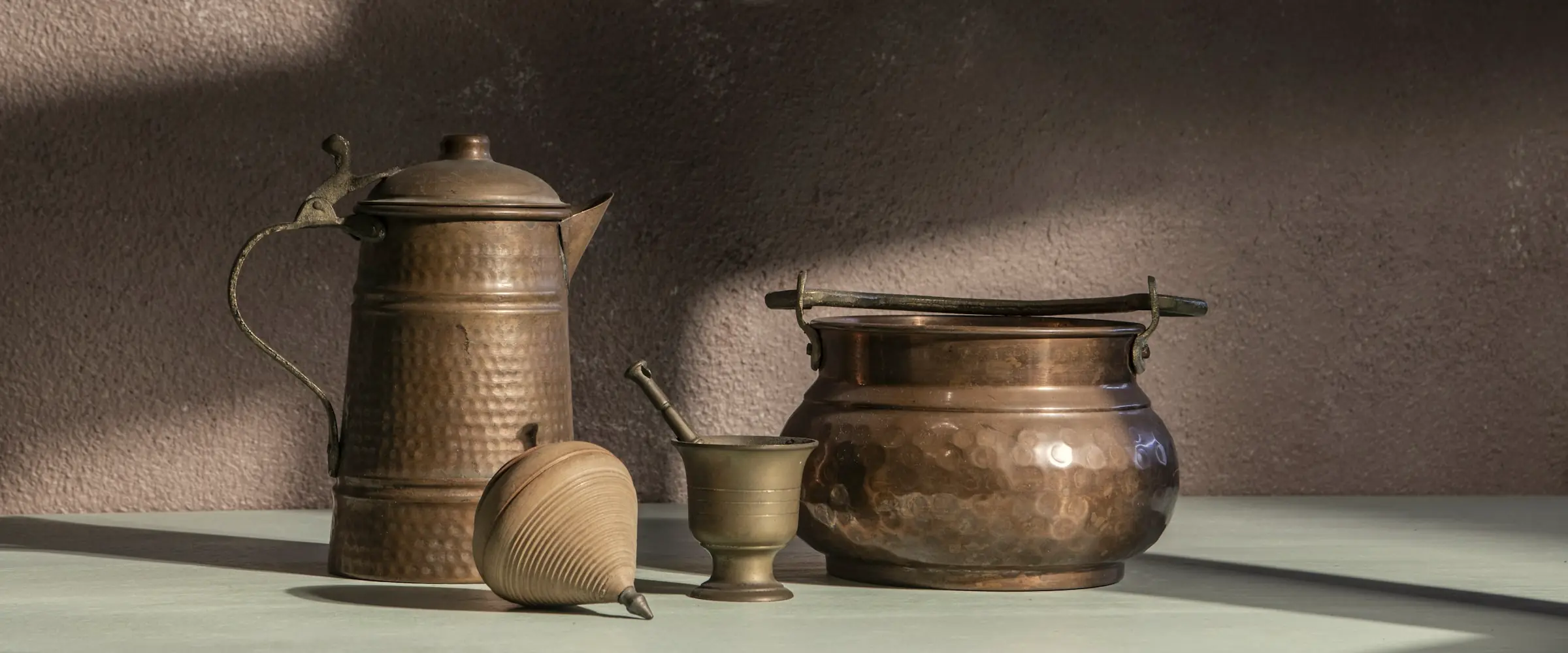 Hammered copper jug, copper pot, brass mortar and a wooden spinning top in low sunlight against a plaster wall
