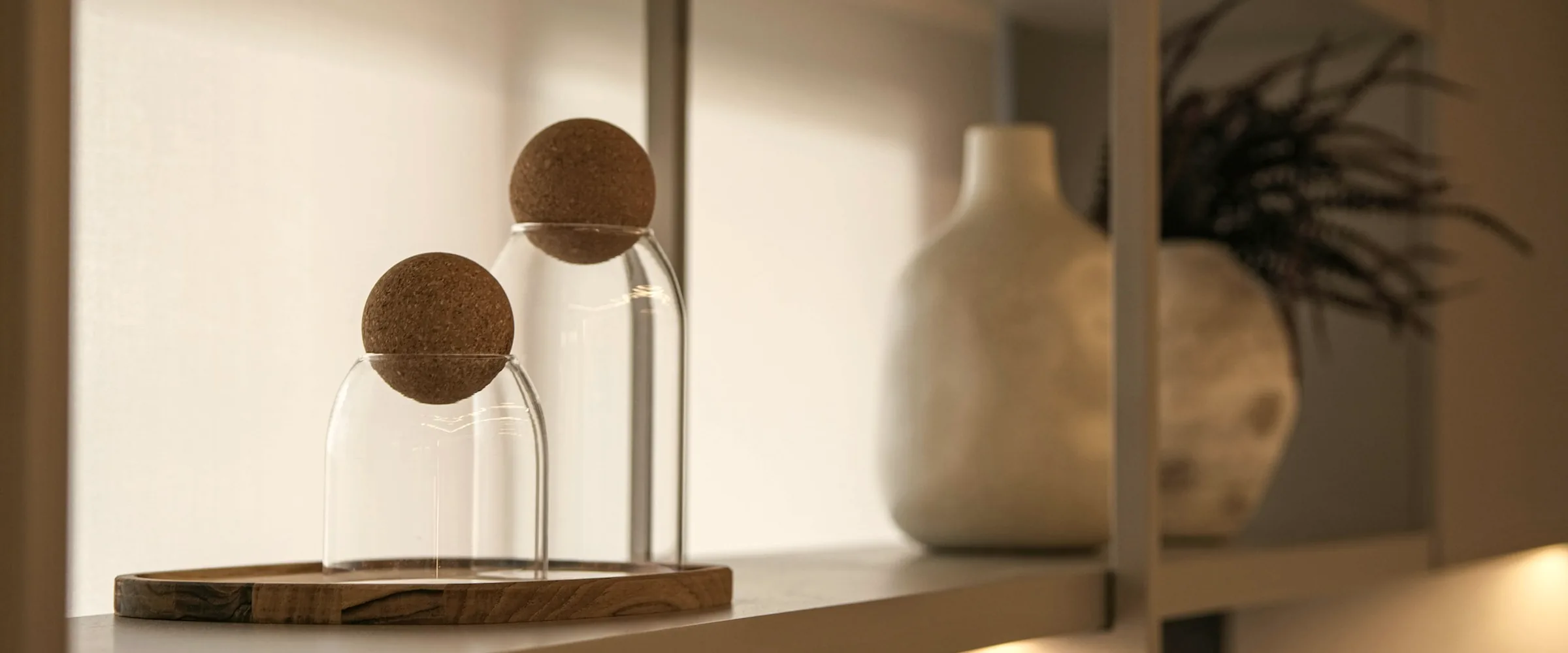 Two glass cloches with cork stoppers on a wooden tray beside a white ceramic vase on a sunlit shelf