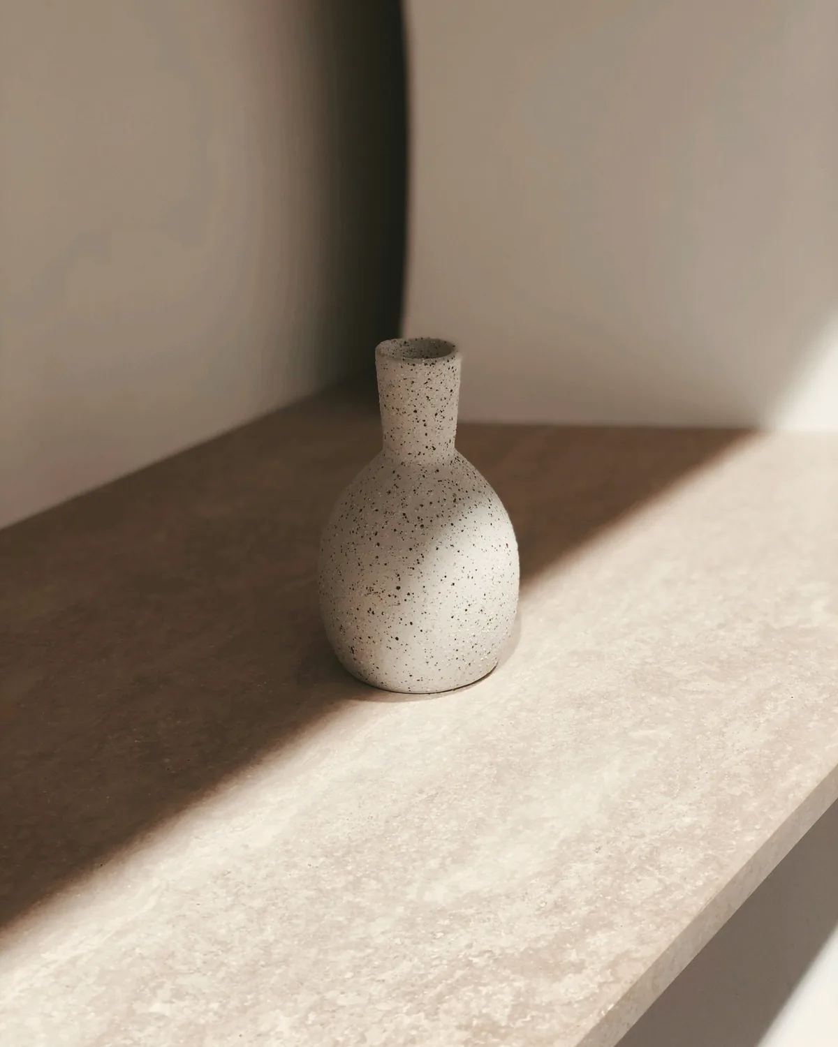 Speckled stoneware bud vase on a travertine shelf in raking sunlight