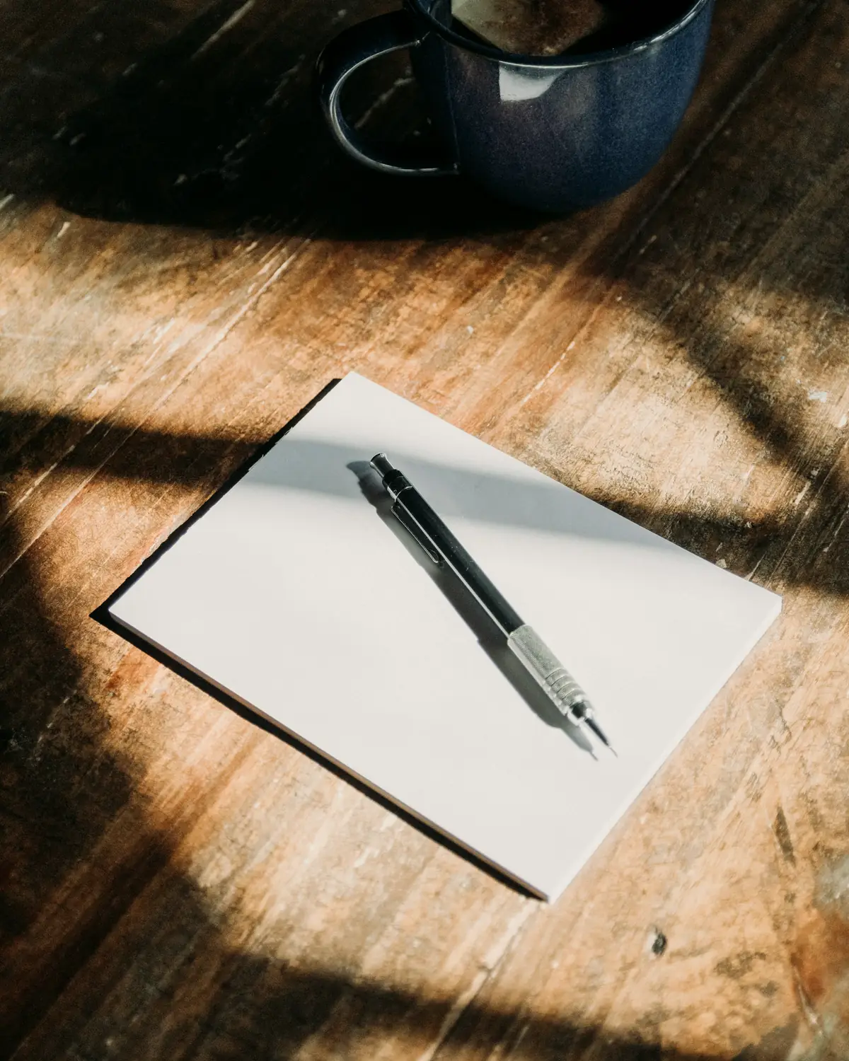 Blank sheet of paper and a pen on a sunlit wooden table beside a cup