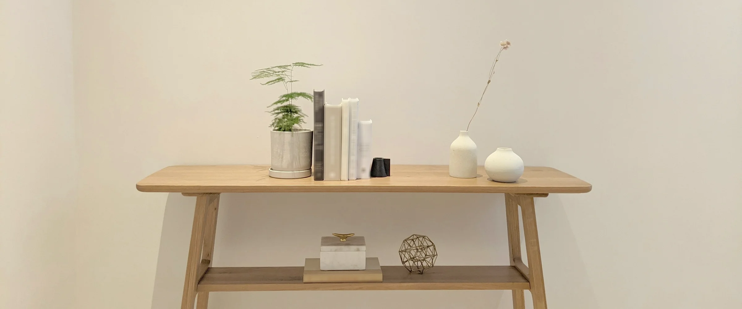 Oak console table with books, a potted fern and two ceramic vases against a pale wall