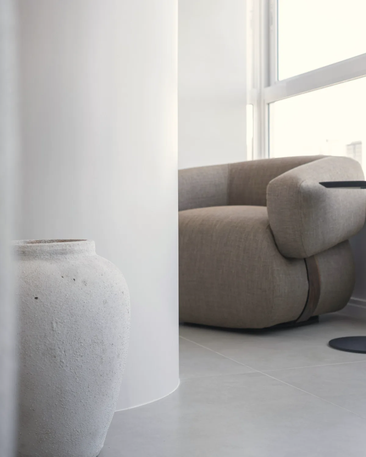 Rounded grey armchair beside a white column and a large textured stone vase in daylight