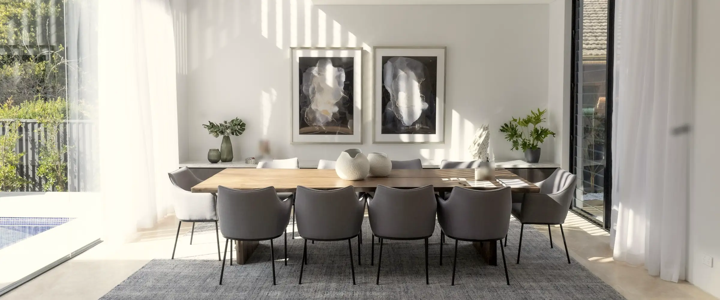 Sunlit villa dining room: a long timber table, grey armchairs and two framed prints above a sideboard
