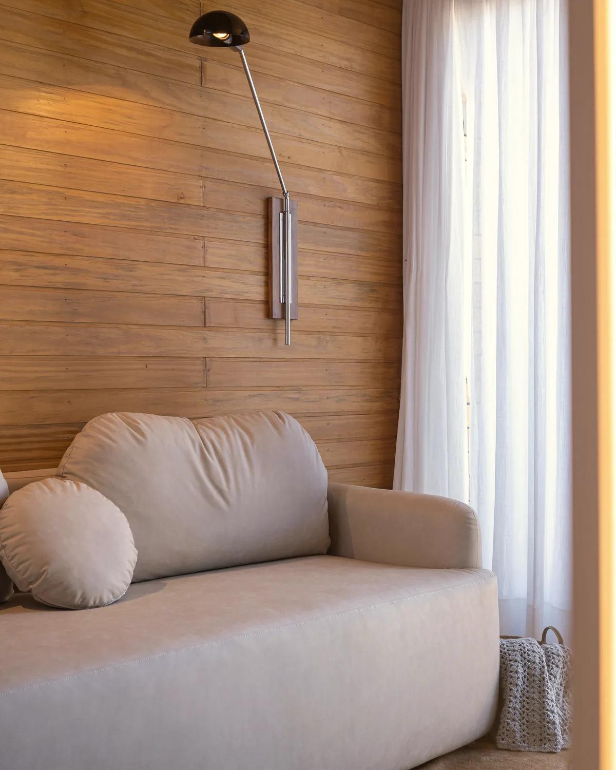 Cream sofa with round cushions against a timber-panelled wall, a swing-arm wall lamp and sheer curtains