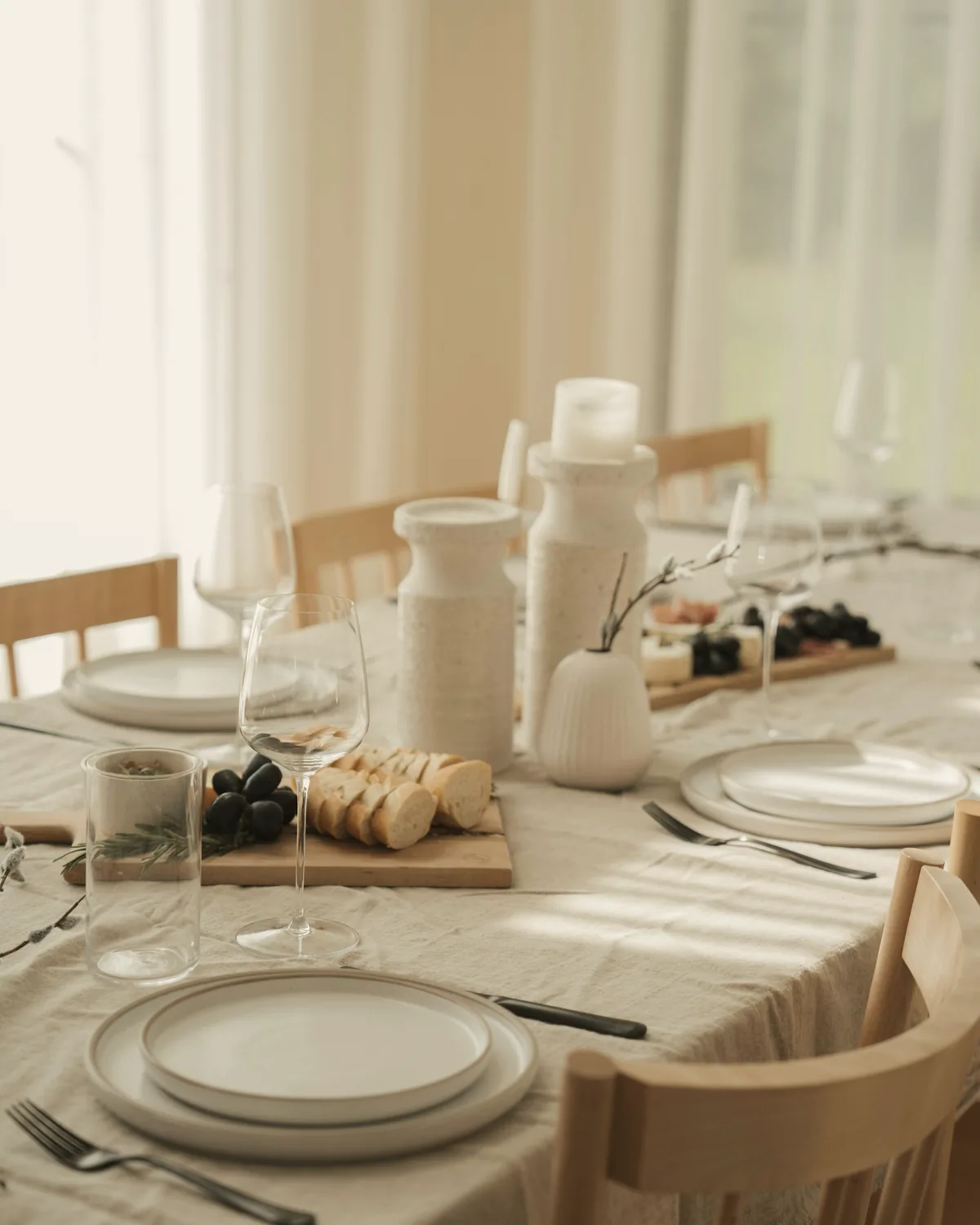A long table laid with linen, white plates, stemware and candles in soft window light