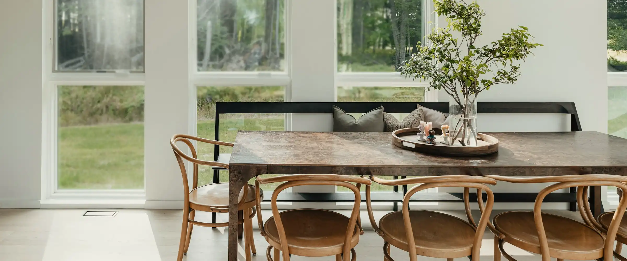 Bentwood chairs around a burl-topped dining table before tall windows, a branch of leaves in a glass vase