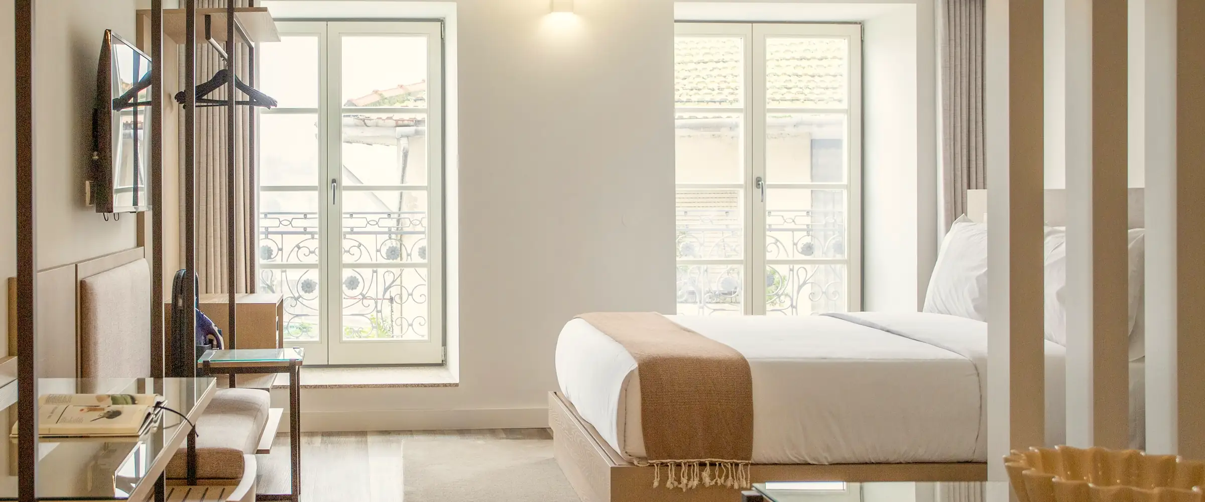 Bright hotel room with oak furniture, a made bed with a woven throw and two tall French windows
