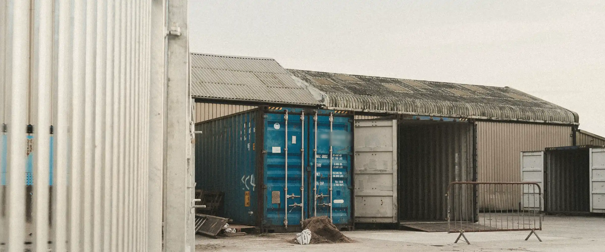A blue shipping container beside an open container and a warehouse in a quiet yard