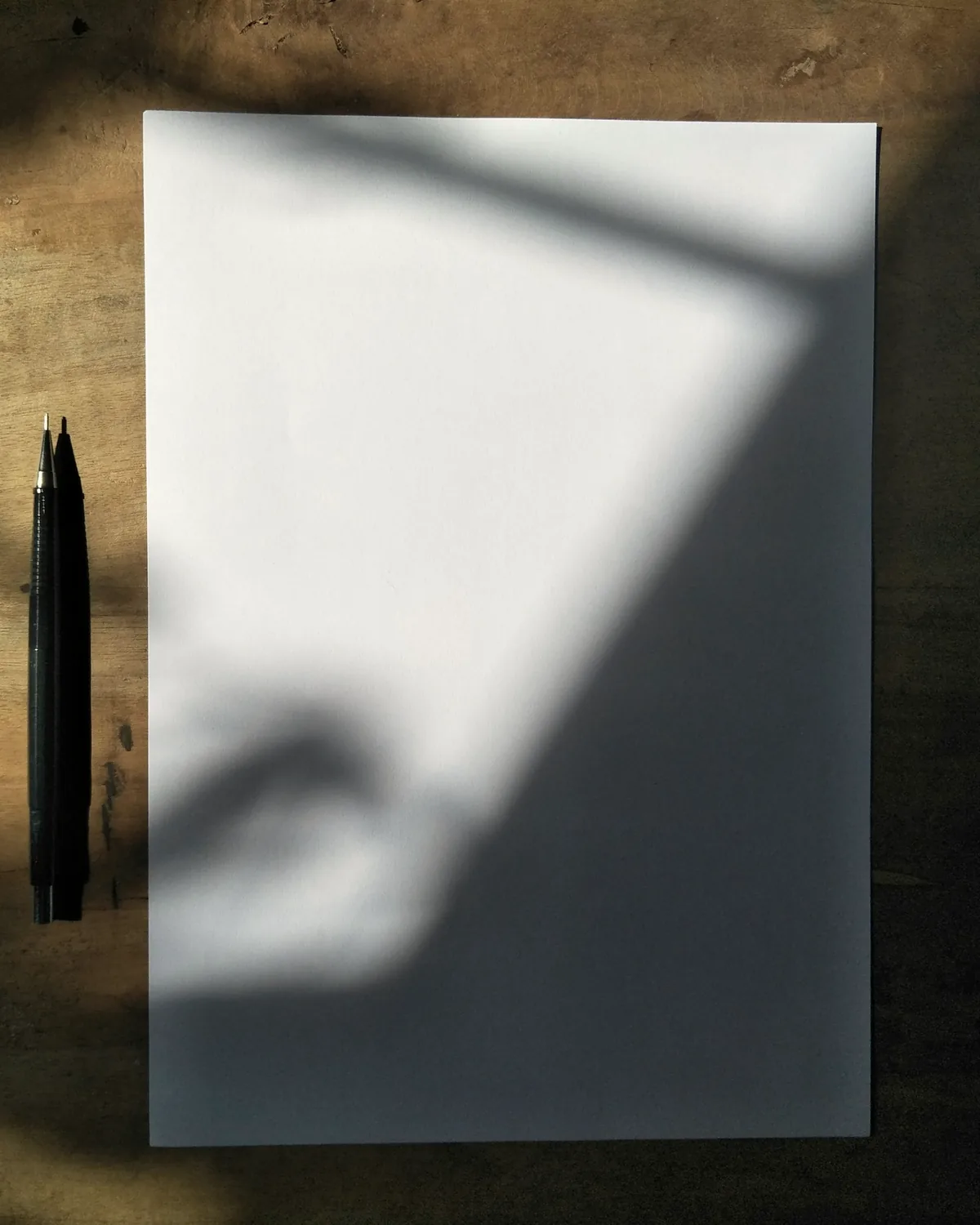 A blank sheet of paper and two pencils on a wooden desk in window light