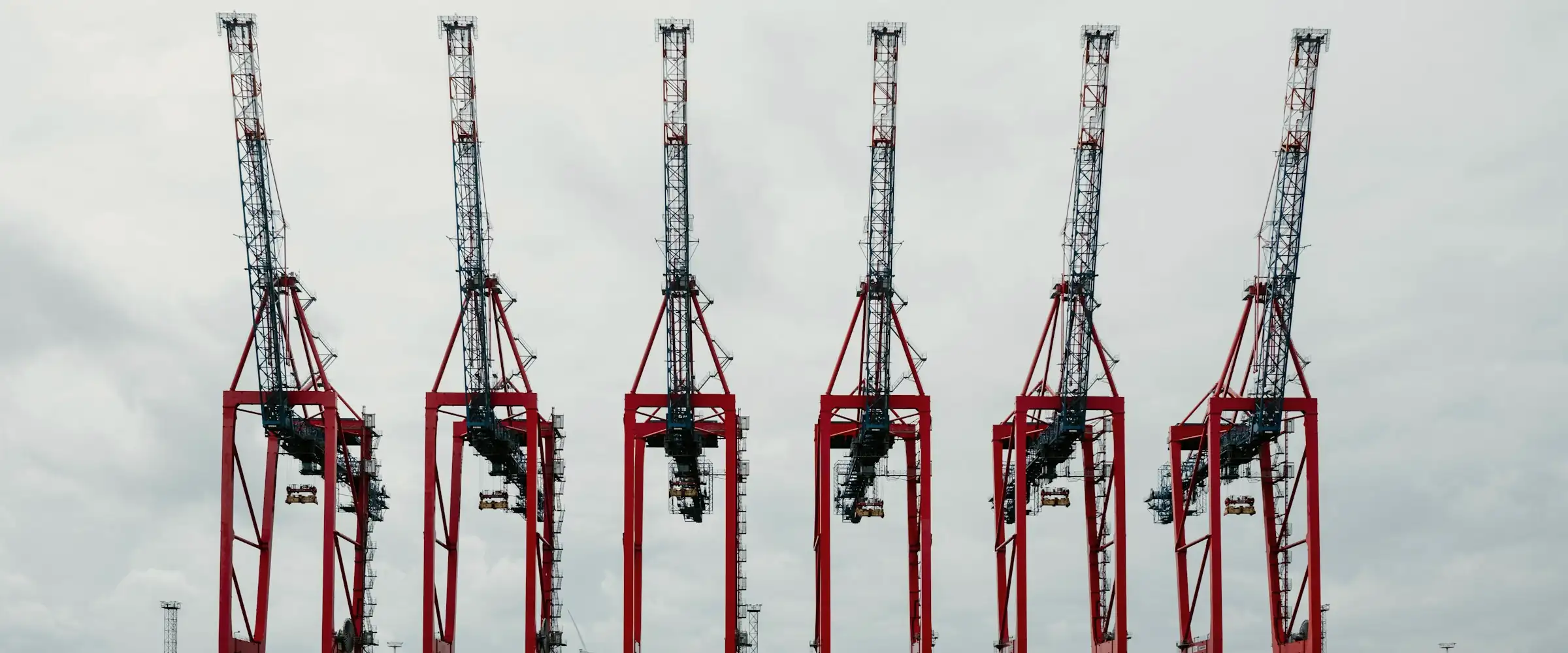 Six red ship-to-shore container cranes in a row against a pale overcast sky