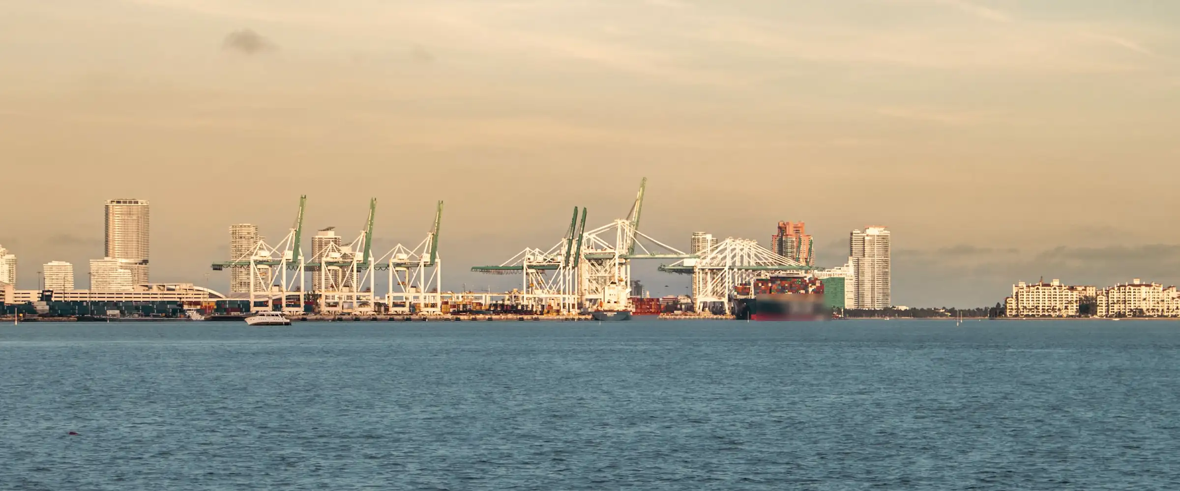 Container port cranes and a city skyline across calm water in warm evening light