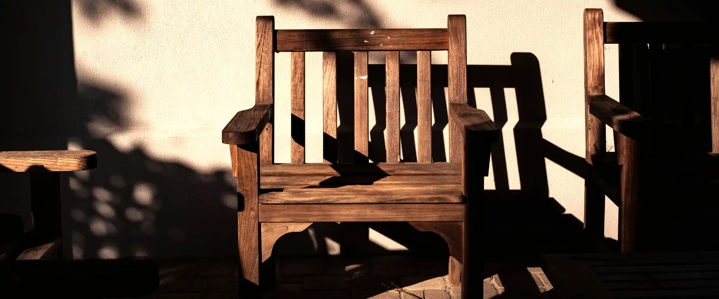 Weathered teak armchair against a pale wall with leaf shadows in late sun