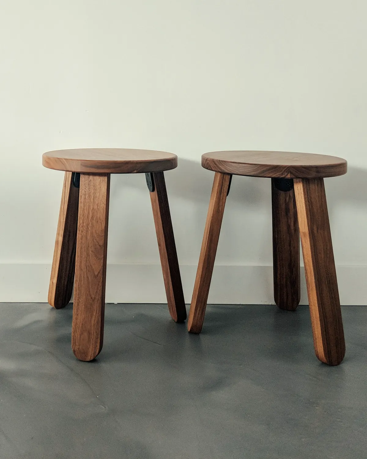 Two walnut three-legged stools against a pale wall on a grey concrete floor