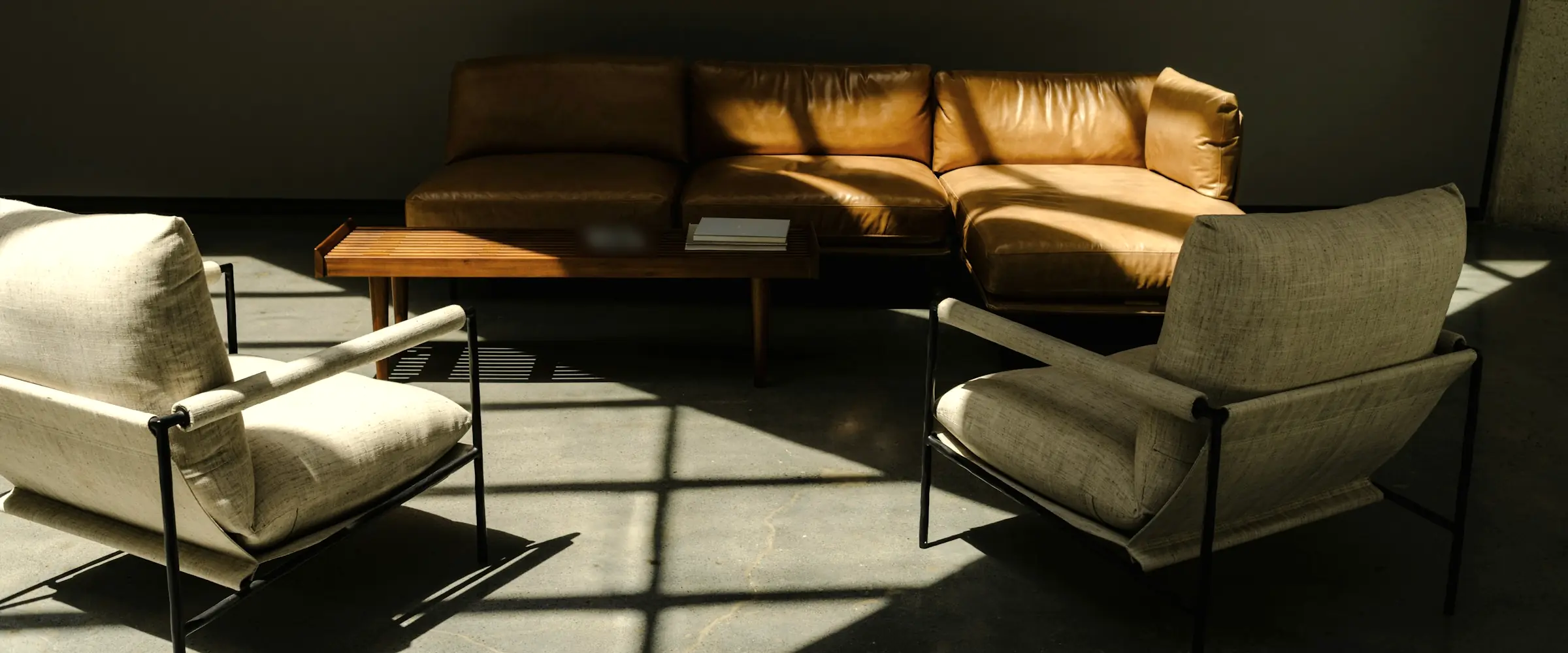 Tan leather sofa, slatted coffee table and two linen lounge chairs in window light on a concrete floor