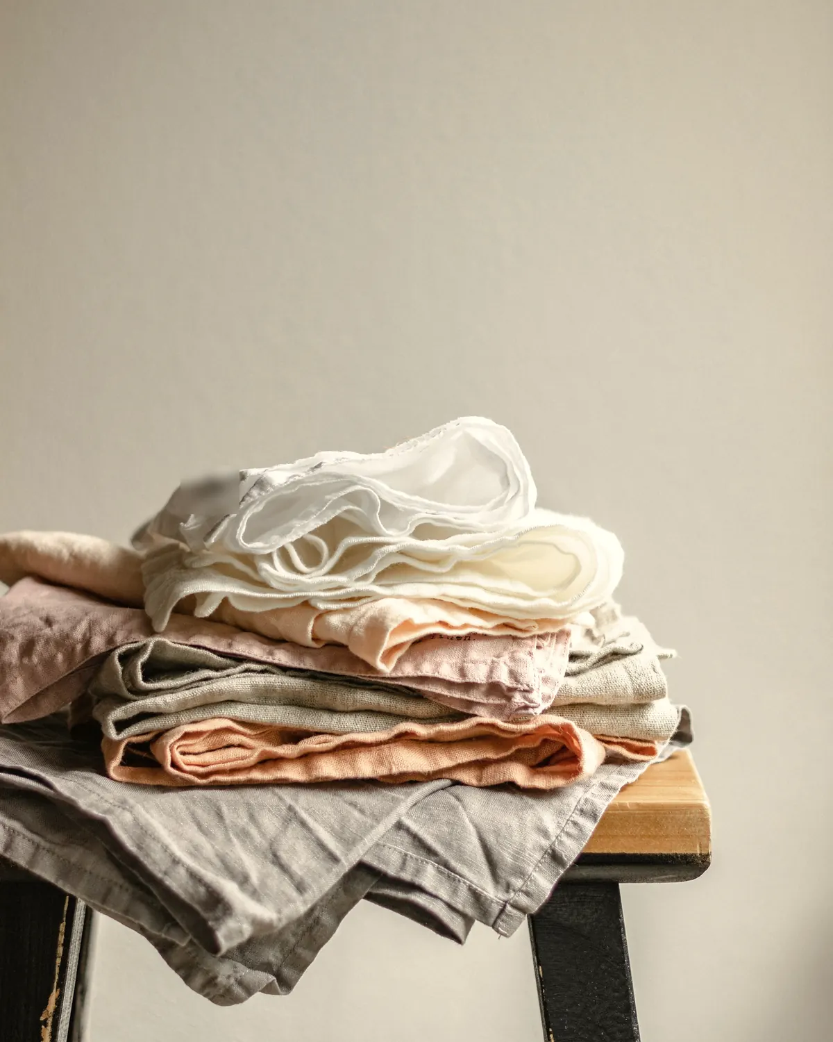 Stacked linen in grey, blush and white on a wooden stool against a plaster wall