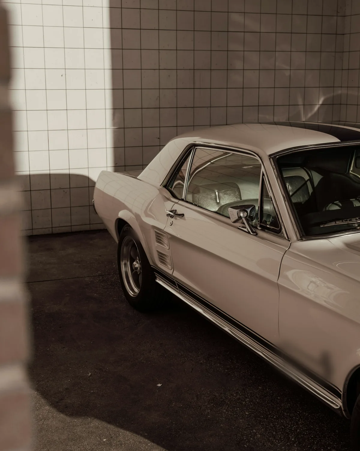 White classic coupe in a tiled garage, sunlight falling across the wall behind it